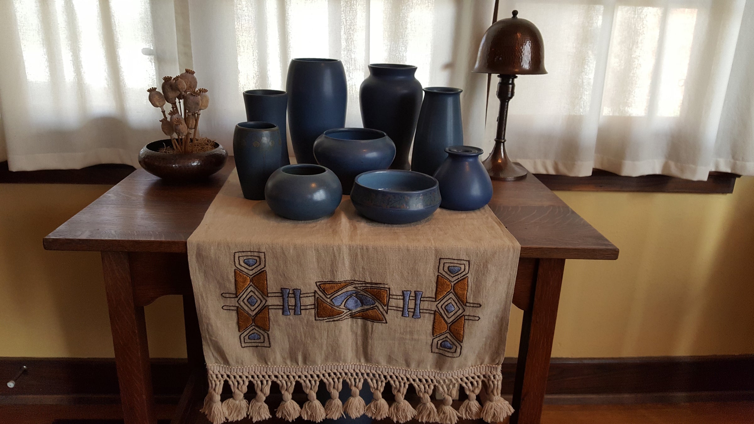 Collection of matte blue studio pottery vases and bowls displayed on an Arts and Crafts oak table with a Southwestern embroidered table runner, complemented by a hammered copper lamp and dried botanicals—perfectly capturing the warm, handcrafted aesthetic of American art pottery interiors
