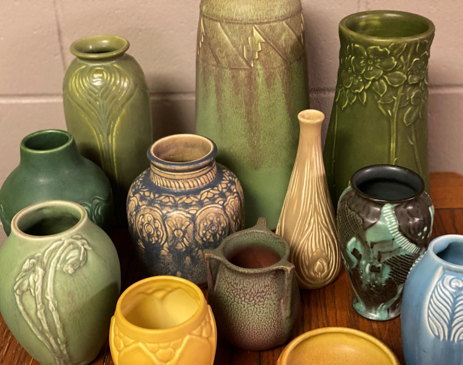 Eclectic collection of vintage art pottery featuring matte and glossy glazes in green, yellow, blue, and earth tones, with decorative relief patterns and handcrafted detailing—highlighting Arts and Crafts style ceramics from makers like Roseville, Weller, and Van Briggle.