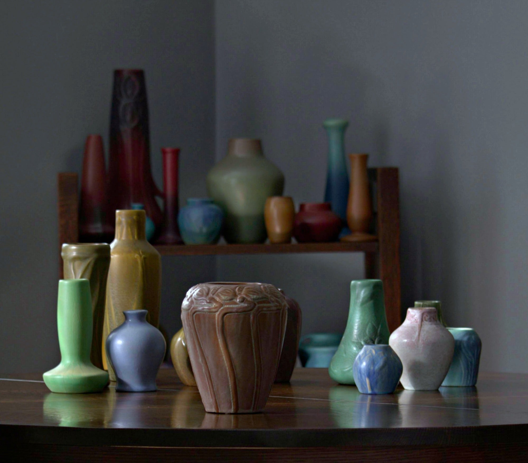 Assorted matte-glazed art pottery vases and vessels in earthy tones of green, ochre, blue, and rose, arranged on a wooden table and shelving unit—highlighting handcrafted forms and rich glazes typical of American Arts and Crafts and mid-century studio pottery.